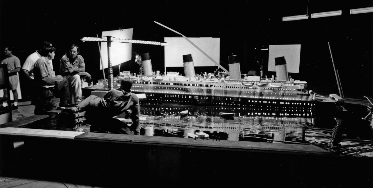 A large model of the ill-fated luxury liner HMS Titanic was set up for lighting tests during prep on the blockbuster period drama Titanic (1997), and at the center of the discussion are (from left) chief lighting technician John Buckley, Carpenter and Cameron (reclining). Of note is the Mylar sheeting around the model, simulating water, which rendered an accurate suggestion of how the huge, to-be-built ship set’s complex practical lighting would play once it was positioned in the tank facility at Fox Baja Studios (seen below).