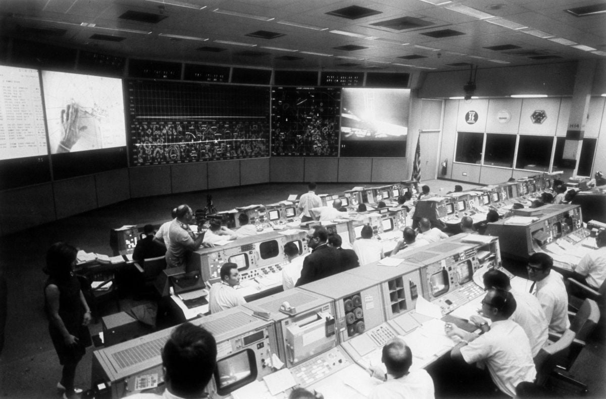 The scene inside the windowless, dimly lit Mission Operations Control Room at NASA’s Manned Spacecraft Center during the Apollo 11 flight. Lead Cameraman Bob Bird of Houston’s A-V Corporation  (standing, at left) moves about filming with his Beaulieu R16B camera, equipped with 9.5-95mm zoom lens. Overhead can be seen banks of mixed blue fluorescent and incandescent lamps with color temperature close to daylight and a light level ranging from 7 to 16 footcandles.