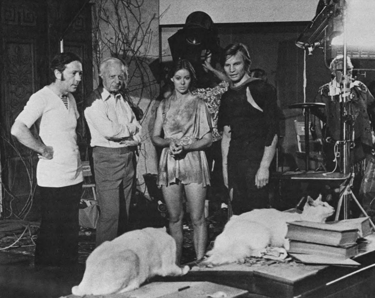 With director Michael Anderson and stars Jenny Agutter and Michael York, the cinematographer checks a couple of cats for photogenic qualities. These are two of the hundreds of cats who appear as the feline friends of Peter Ustinov in a sequence that takes place in a vine-overgrown set representing the U.S. Senate chamber 300 years in the future.