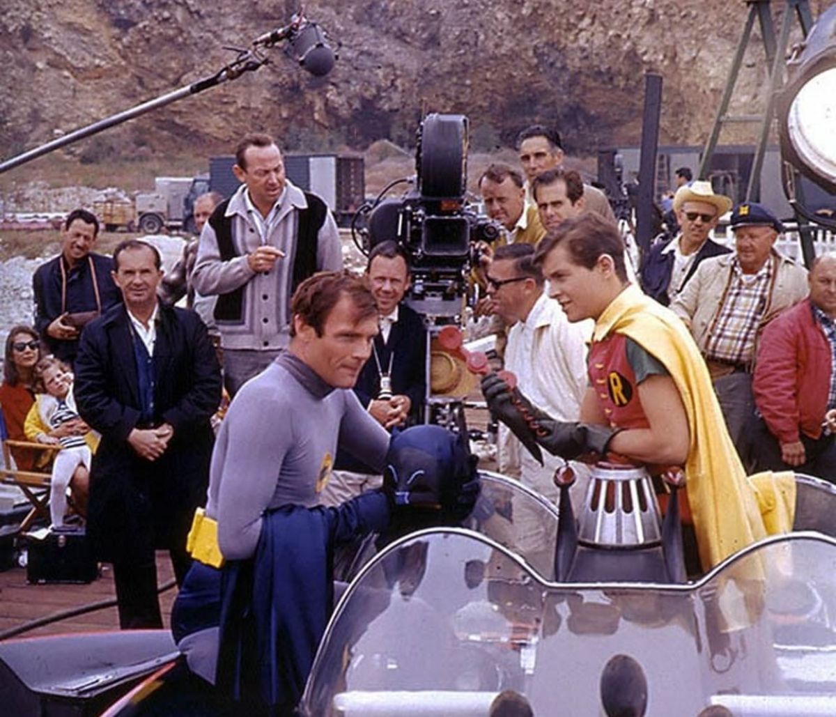 Cast and crew from 20th Century-Fox Studios on location filming a feature-length version of Batman, nation’s most popular TV series. Batman (Adam West) and Robin (Burt Ward) are shown in foreground rehearsing a scene with Batmobile and Batphone, while Director Les Martinson (under camera) and Director of Photography Howard Schwartz, ASC (in striped-V sweater), check the action.