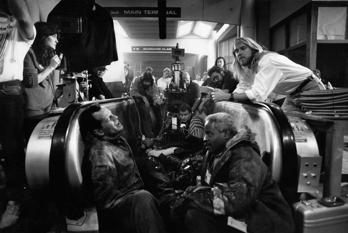 Three cameras line up for coverage on co-stars Willis and Art Evens as director by Renny Harlin stands by (observing from right). Most of the film’s interior airport scenes were filmed in the Tom Bradley International Terminal at Los Angeles International Airport. Wood can be seen in the background directly behind the center camera, wearing a white shirt, his head slightly tilted as he watches the scene.